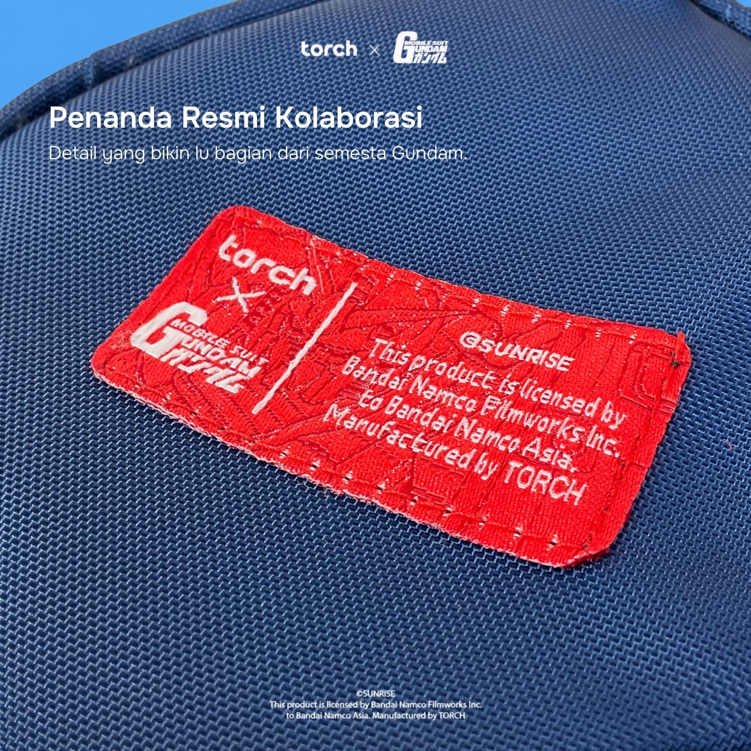 A close-up of a blue woven fabric surface with a prominent red embroidered patch. The patch includes white text reading 'torch x GUNDAM' and additional licensing text, with white overlaid text at the top mentioning a collaboration.