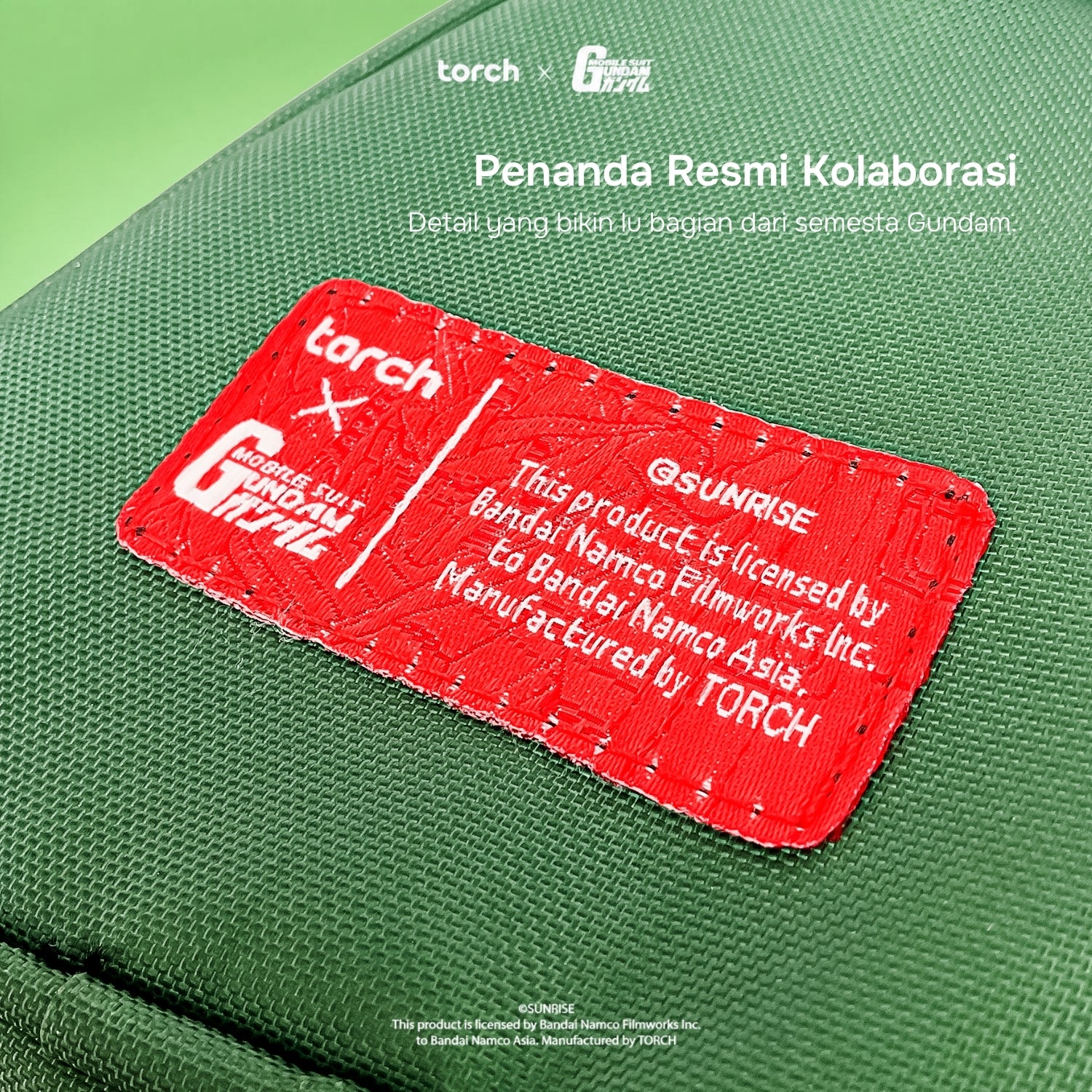 A close-up of a green fabric pouch or bag with a prominent red stitched label. The label has white text reading 'torch x Gundam' and additional licensing text, with white overlaid text at the top reading 'Penanda Resmi Kolaborasi' and small 'torch G-DRIVE' marks.