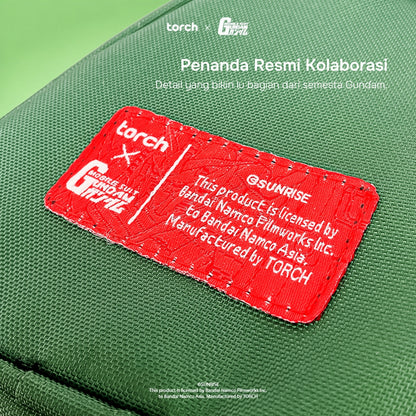 A close-up of a green fabric pouch or bag with a prominent red stitched label. The label has white text reading 'torch x Gundam' and additional licensing text, with white overlaid text at the top reading 'Penanda Resmi Kolaborasi' and small 'torch G-DRIVE' marks.