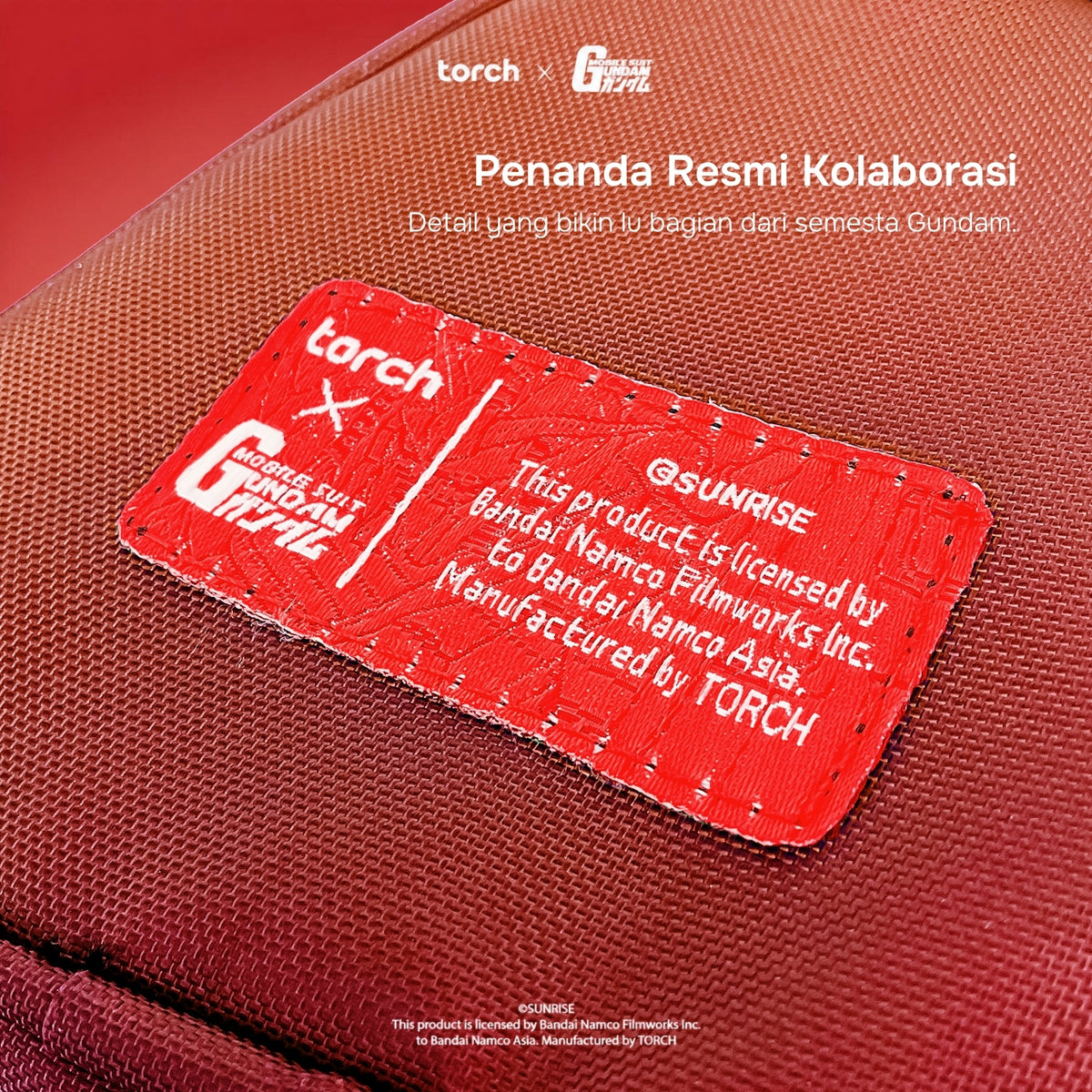 Close-up of a textured red fabric pouch or bag with a sewn red label reading 'torch x Gundam' and additional white text at the top about official collaboration details.